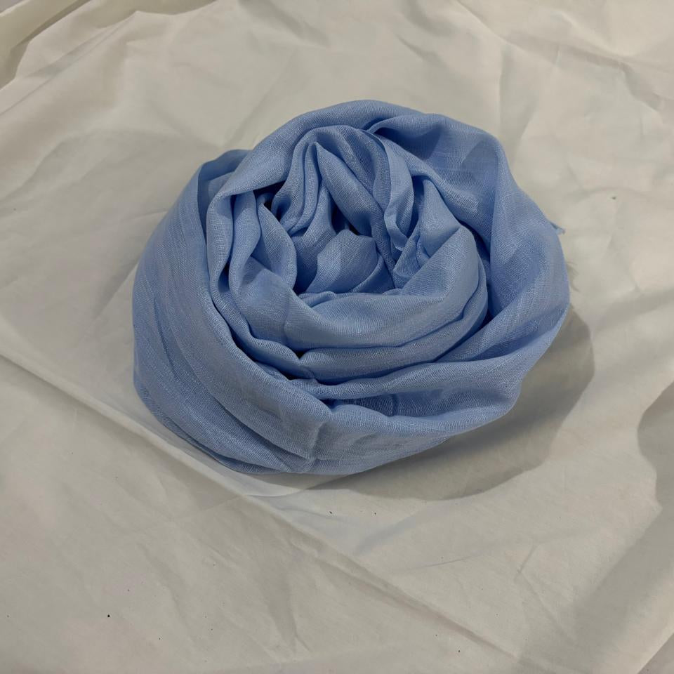 A light blue scarf or shawl, neatly rolled into a spiral, displayed on a wrinkled white fabric background.