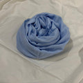 A light blue scarf or shawl, neatly rolled into a spiral, displayed on a wrinkled white fabric background.