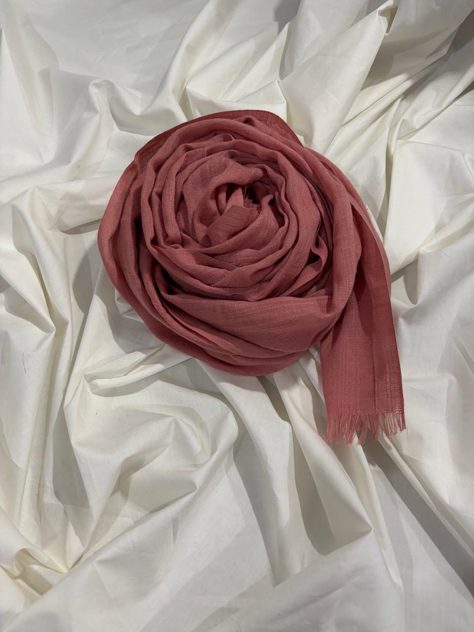 A dusty rose pink scarf, neatly rolled and draped on a background of rumpled white fabric.