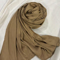 A draped, matte, opaque scarf in a warm gold shade, arranged in a loose spiral on a wrinkled white fabric background.